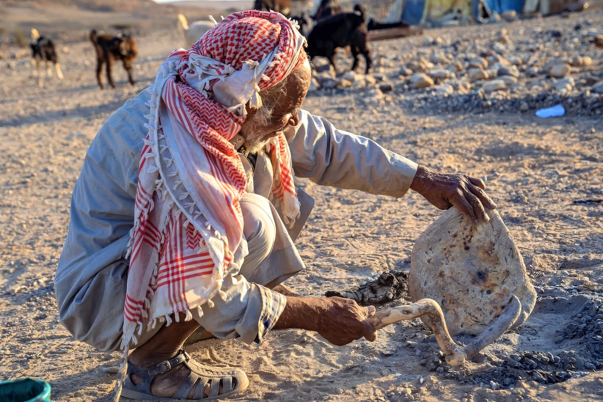 A Bedouin is baking bread in the desert near his home in Jordan. He's dusting it off with a stick after baking it on a fire in the sand.