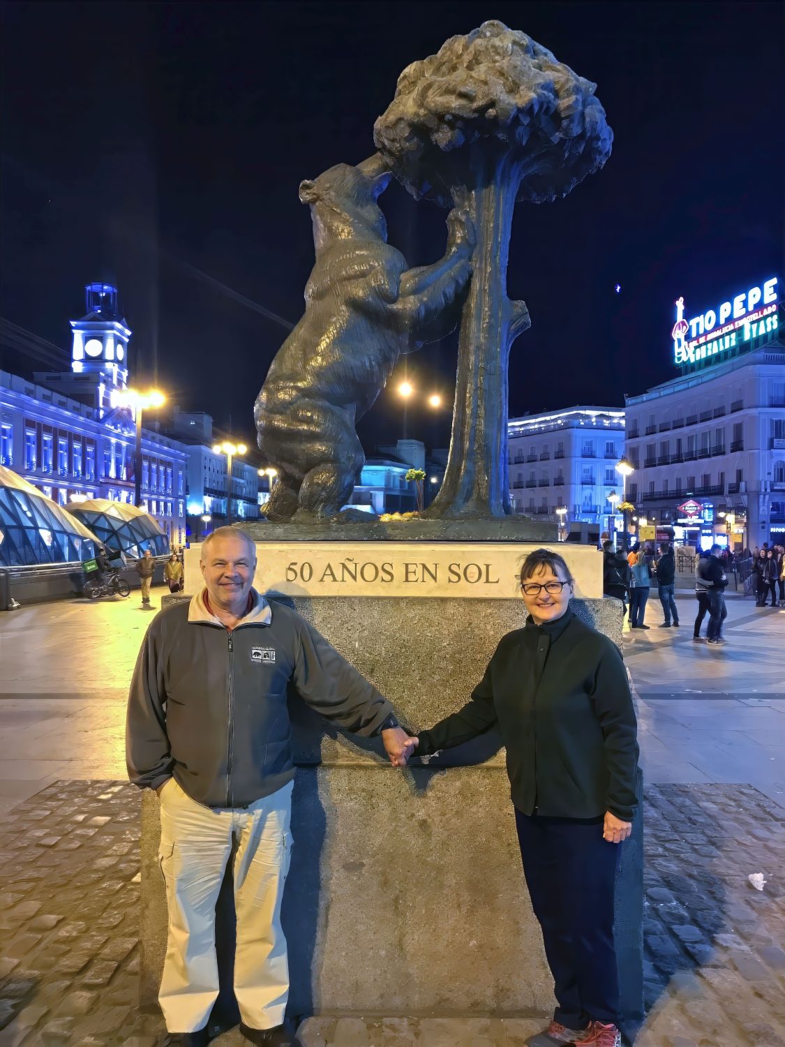 Marie and Eddie are holding hands in front of the Statue of the Bear and Strawberry Tree in Madrid's historic city center.