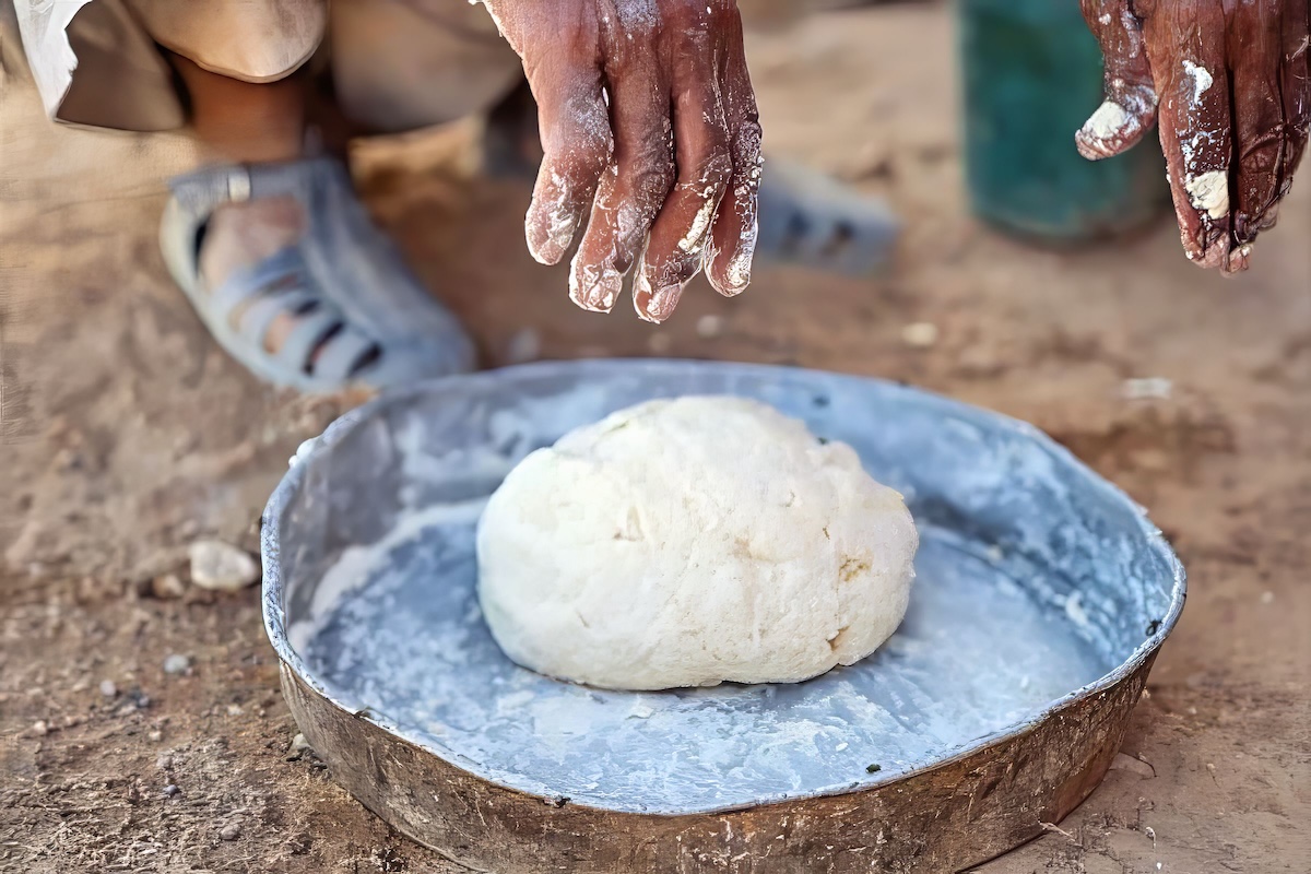 A Bedouin is squatting and baking bread, with his finger hanging down towards the ball of dough in the pan on the ground. 